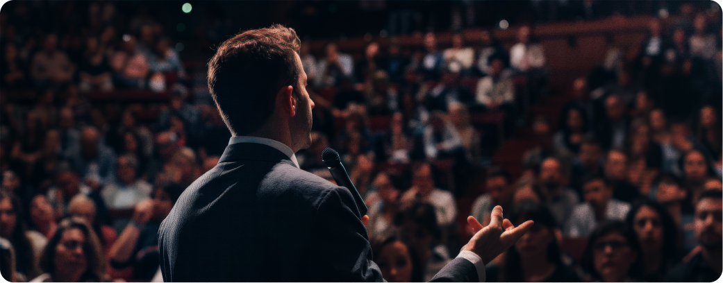 Homem palestrante em palco em frente a uma plateia cheia, com o braço estendido