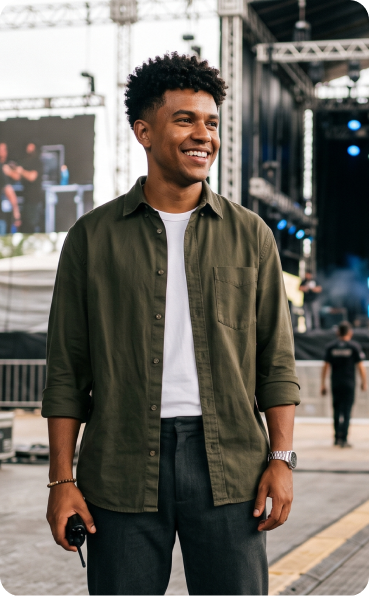 Jovem sorridente segurando rádio comunicador em frente a um palco de festival, vestindo camisa verde.