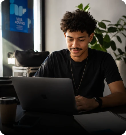 Jovem utilizando um notebook para gerenciar atividades em uma mesa com copo de café, em um ambiente de escritório.