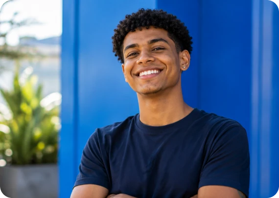 Jovem sorridente com os braços cruzados vestindo camiseta escura em frente a uma estrutura azul brilhante.
