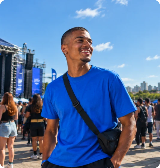 Jovem sorridente usando camiseta azul e bolsa transversal aproveitando um evento diurno ao ar livre.